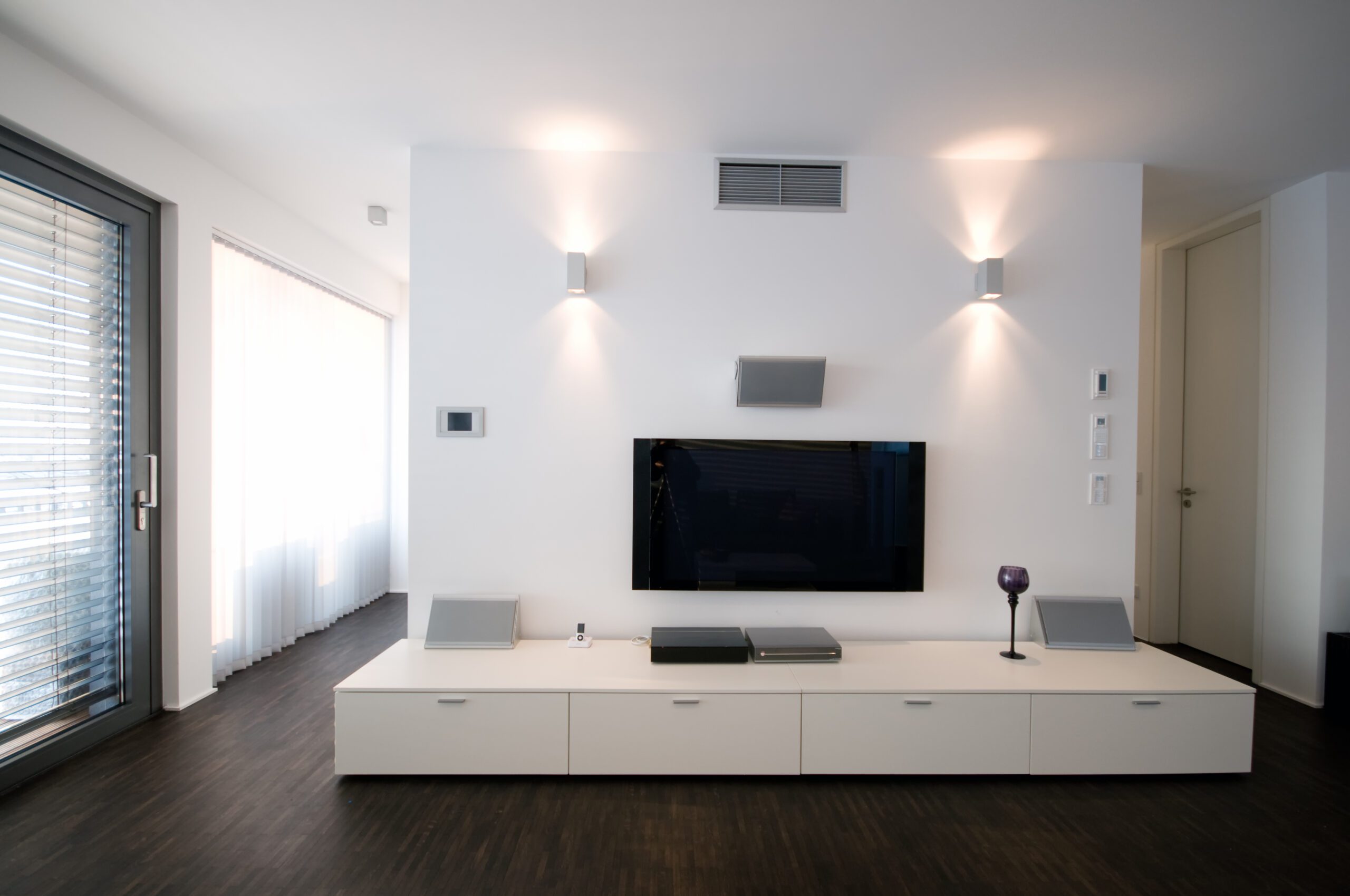 Modern,Home,Theater,Interior A modern living room features a wall-mounted flat-screen TV with a minimalist white media console below, illuminated by two wall-mounted lights. Vertical blinds cover a large window beside them.